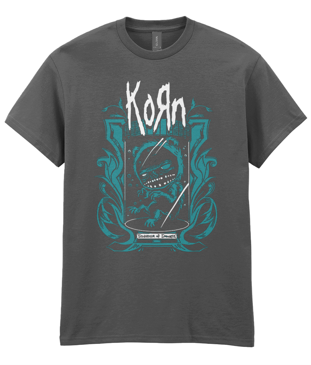 A dark gray short-sleeve t-shirt featuring a graphic print of the band Korn. The design includes the band's name in a stylized white font above a teal and white illustration of a lizard inside a shield, with a 'Welcome to the Underworld' banner at the bottom.